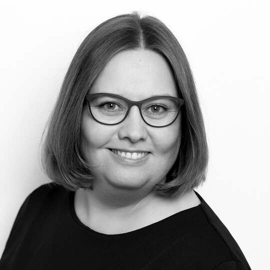 Porträt von Katrin Floß. Sie trägt eine dunkle Brille und ein schwarzes Oberteil. Ihr braunes, kinnlanges Haar ist glatt gestylt. Sie lächelt in die Kamera vor einem hellen Hintergrund.