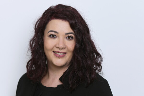 Porträt von Angelique Lobjinski. Sie hat schulterlanges, dunkel gelocktes Haar und trägt ein schwarzes Oberteil. Sie lächelt freundlich in die Kamera. Sichtbar sind ein Nasen- und ein Lippenpiercing. Der Hintergrund ist hell.