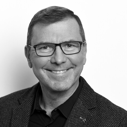 Porträt von Peter Eichler. Er trägt eine Brille, ein dunkles Hemd mit gemusterter Jacke und lächelt in die Kamera. Der Hintergrund ist hell und neutral.