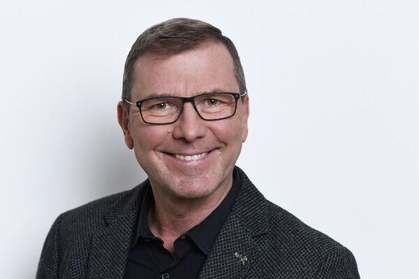 Porträt von Peter Eichler. Er trägt eine Brille, ein dunkles Hemd mit gemusterter Jacke und lächelt in die Kamera. Der Hintergrund ist hell und neutral.