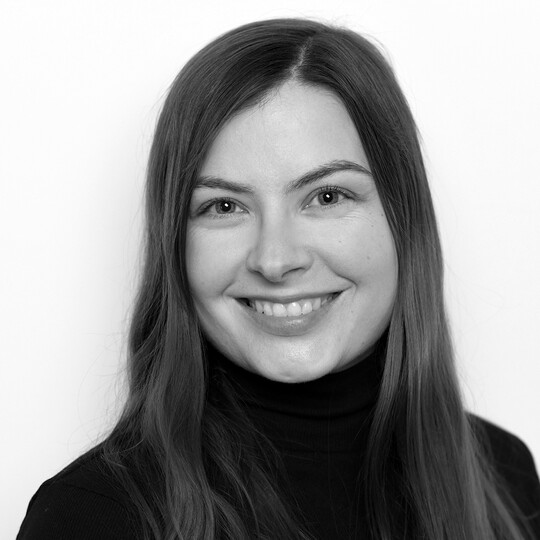 Porträt von Romy Kleinert. Sie hat langes, glattes, braunes Haar und trägt ein schwarzes Oberteil mit Rollkragen. Sie lächelt freundlich in die Kamera, vor einem hellen Hintergrund.