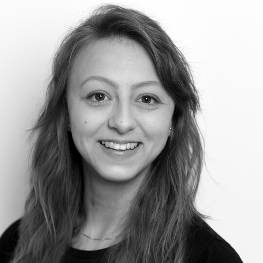 Porträt von Nina Hahn. Sie trägt ein dunkles Oberteil und hat lange, wellige, hellbraune Haare. Sie lächelt in die Kamera, im Hintergrund eine helle Wand.