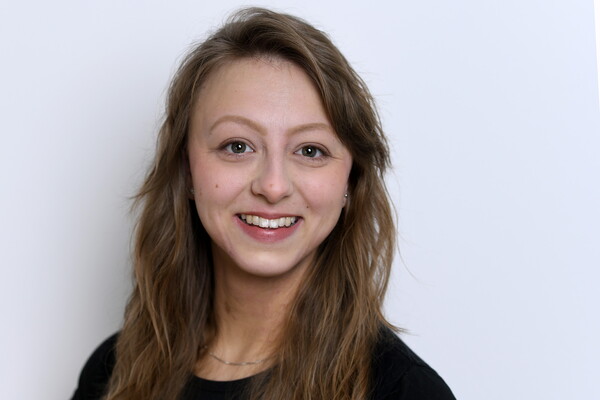 Porträt von Nina Hahn. Sie trägt ein dunkles Oberteil und hat lange, wellige, hellbraune Haare. Sie lächelt in die Kamera, im Hintergrund eine helle Wand.