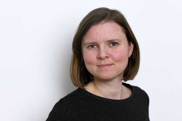 Porträt von Carolin Heckel. Sie hat kinnlanges, glattes, braunes Haar und trägt ein dunkles Oberteil. Sie blickt ruhig und freundlich in die Kamera, vor einem hellen Hintergrund.